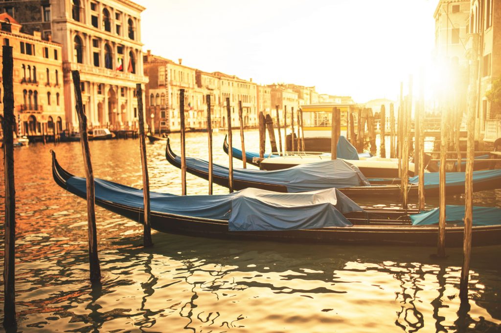 Gondolas lined up in a canal in Venice, with the sun bright and golden covering the buildings in an orange glow.