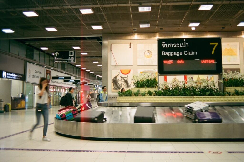 People collecting luggage at an airport baggage claim area marked with a sign in both English and Thai, hoping not to experience lost luggage.