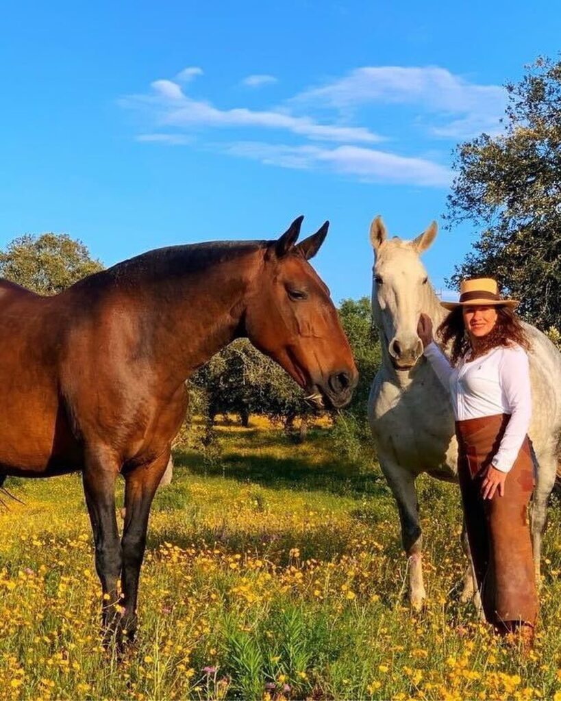 A woman in a hat stands in a field with two horses, one brown and one white, amid yellow flowers under a clear sky in local Portugal.