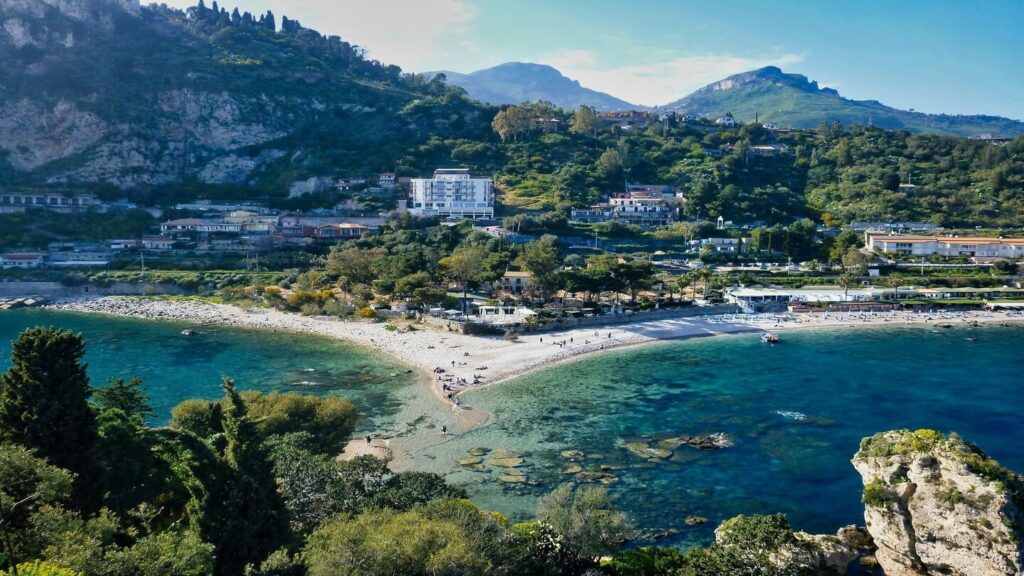 A scenic view of a coastal village with a curving beach, clear blue water, and lush green mountains in the background highlights current vacation trends.