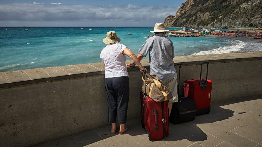 Two older adults with suitcases facing the sea, one in a white hat, leaning on a seafront balustrade under a partly cloudy sky.