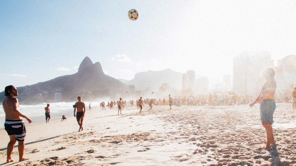 People play football on a sunny beach with the best cities to visit in September and mountains in the background.