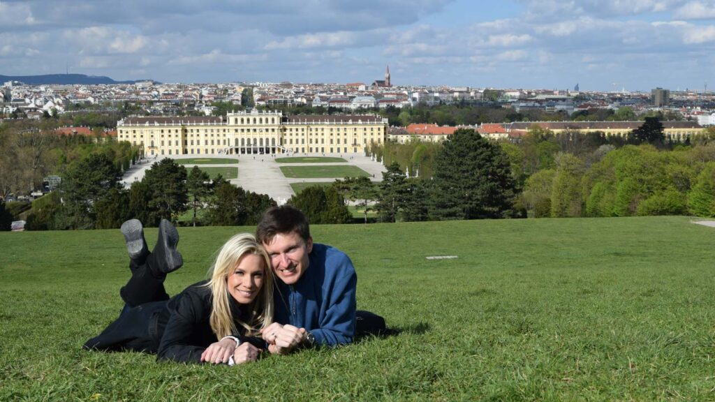 Ashlinn and her husband lying on a grassy hill in Vienna, Austria with the Schönbrunn Palace in the background