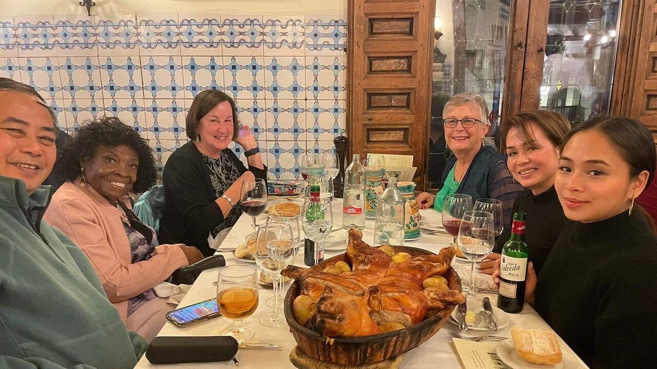 A group of people at a dinner table smiling for a photo, tiles are in the background and a large pork dish sits in the middle