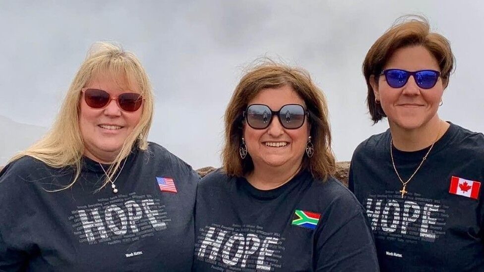 Three friends pose for a photo with 'Hope' T-shirts on and small flags pinned to the shirts, displaying the country that they're each from. One from USA, one from South Africa, one from Canada