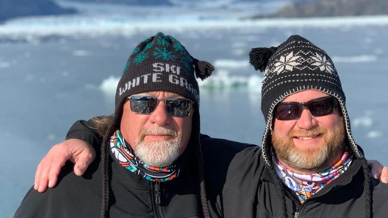 Father and Son in fleece, snow hats and sunglasses posing for a photo with Alaskan glacier in background. Great travel stories made with dad.