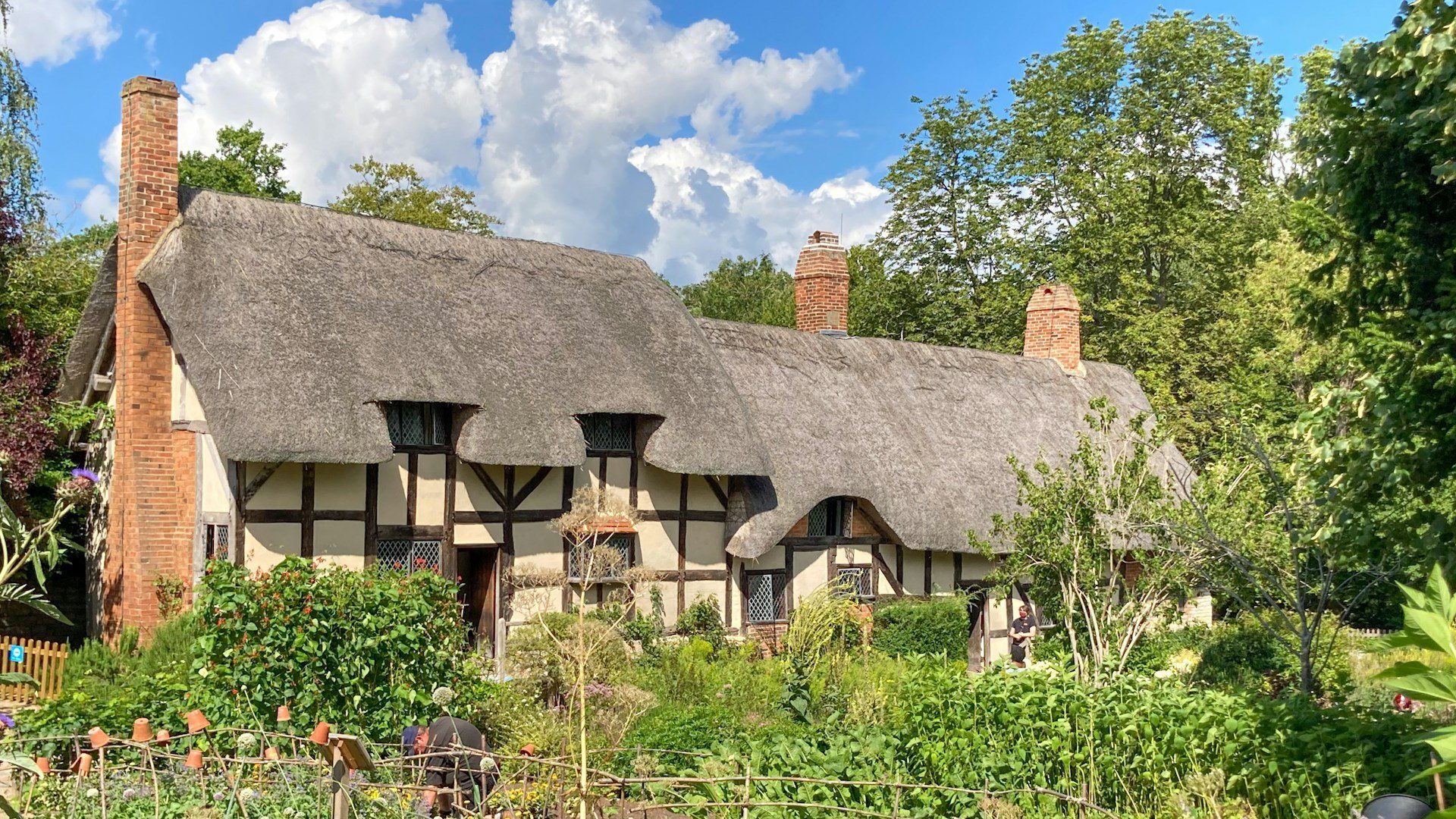 Photo of a large thatched cottage, with three brick chimney stacks, surrounded by leafy trees and gardens