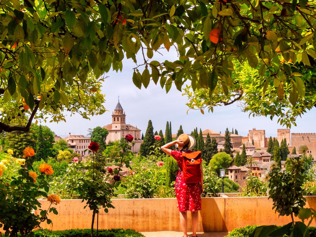 A woman in a red dress and straw sunhat wearing an orange backpack photographed from behind looking out over buildings in a European city