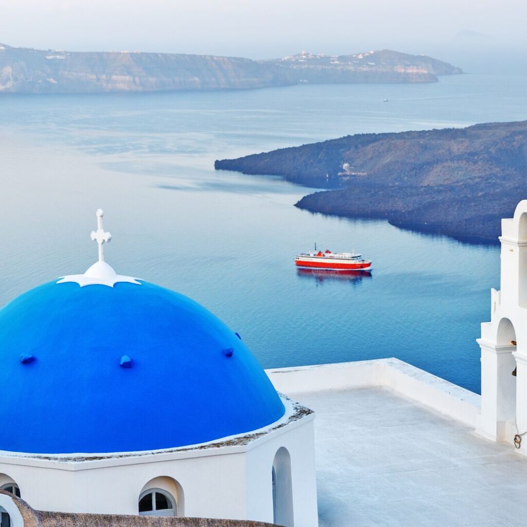 A blue dome of a building in Santorini is in the foreground, while beautiful islands are in the background. A red cruise ship is sailing between the land