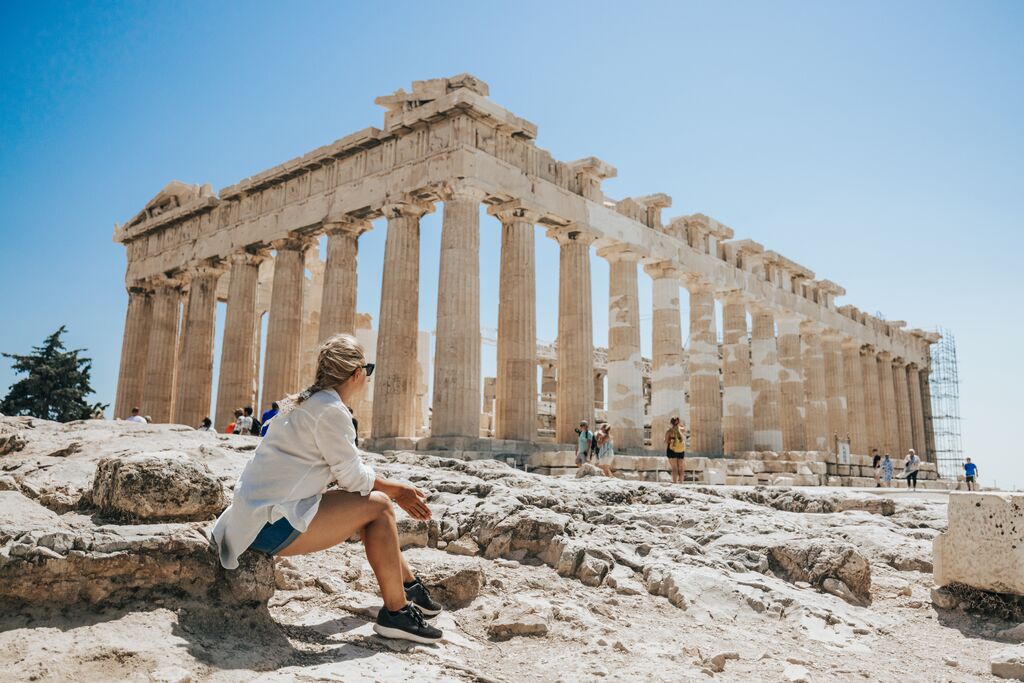 A woman sits while staring at a beautiful Greek ruin, huge columns standing up in a rectangle shape.