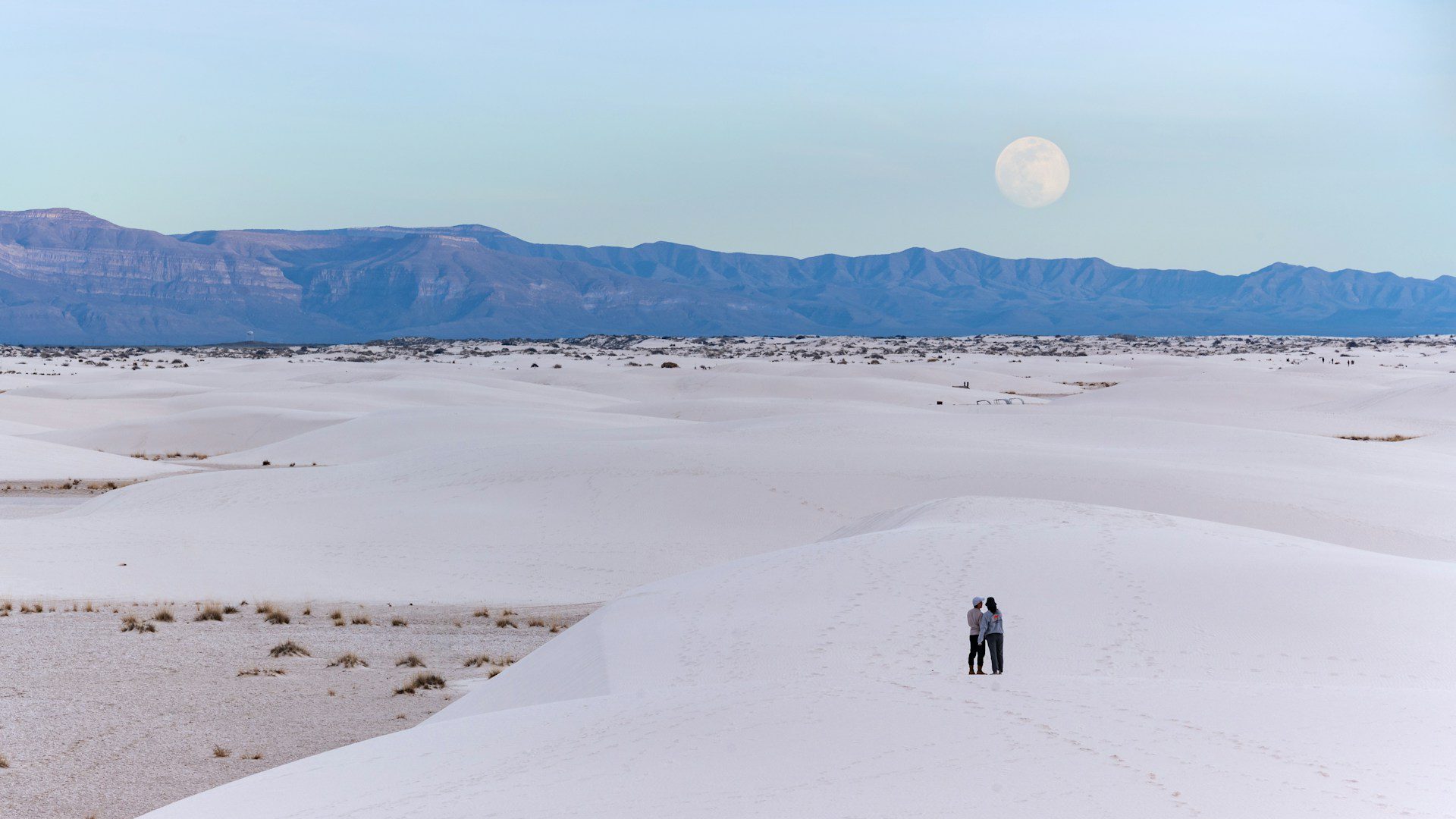 Photo of a couple standing among the white sand dunes, with mountains and a full moon in the distance