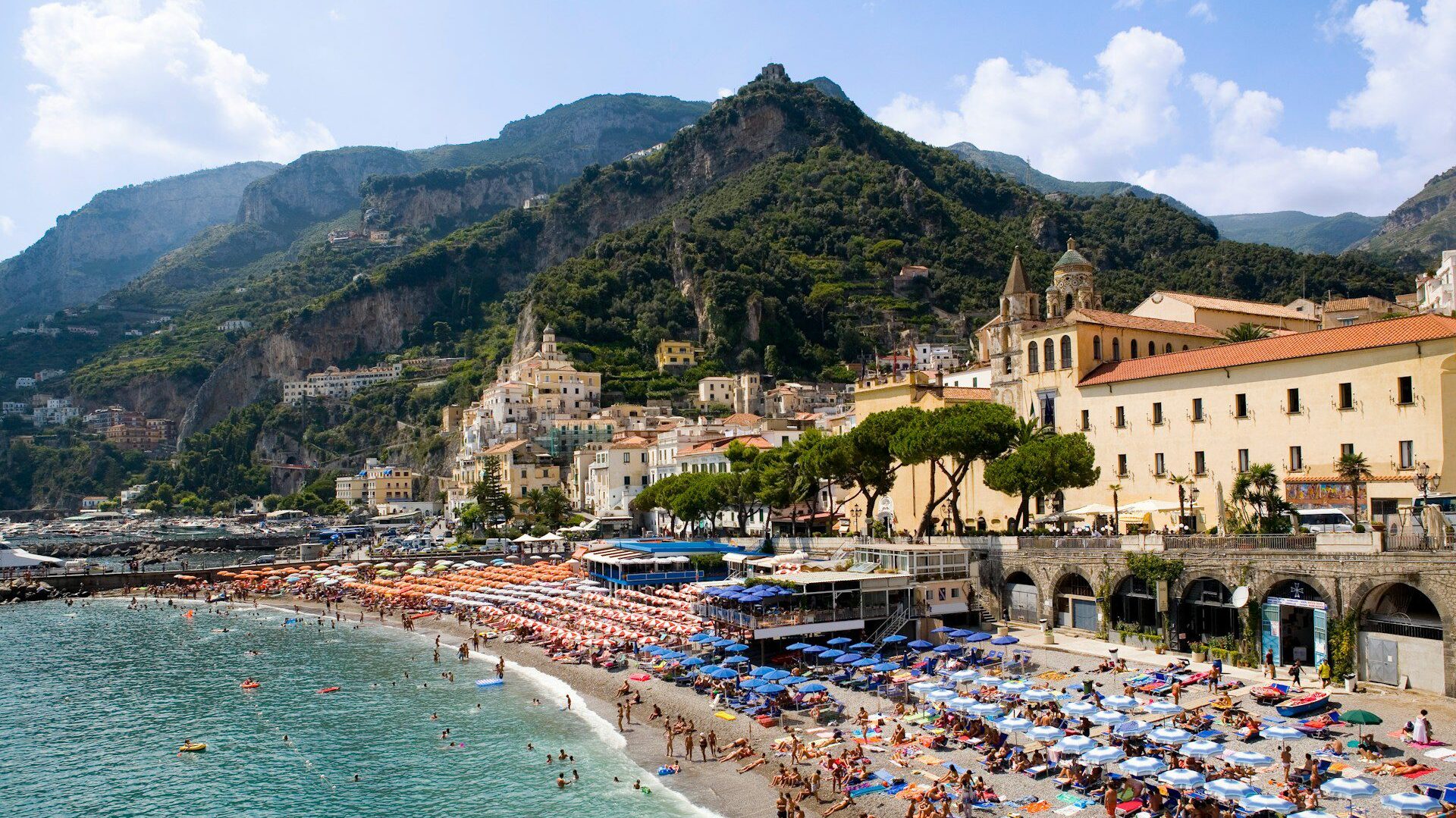 A beach and sea front with jagged  green hills in the background and turquoise water